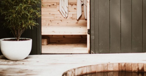 A modern outdoor sauna with cedar wood interior, black exterior paneling, a white potted plant, and a wooden hot tub.