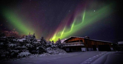 a large alaska cabin covered in snow with surrounding wintery trees and the northern lights visible in the sky