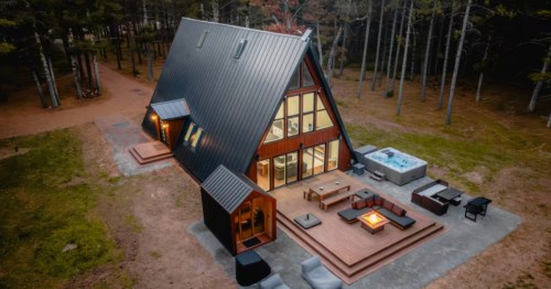 An aerial view shows a modern A-frame cabin in a wooded setting, featuring a large deck with a fire pit, outdoor seating, and a hot tub.