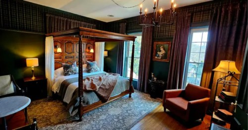 A luxurious bedroom features a dark wood, four-poster bed with plush bedding and decorative pillows. A chandelier hangs above, and the walls are dark with patterned wallpaper. Two windows flank the bed, and seating is arranged in the room.