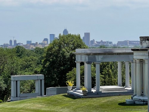 A scenic view of Crown Hill Cemetery in Indianapolis, Indiana, featuring classical-style mausoleums and monuments in the foreground. The lush greenery of the cemetery contrasts with the modern skyline of downtown Indianapolis in the background, including the city's tallest buildings. The overcast sky provides soft, diffused lighting, enhancing the serene atmosphere of the historic site.