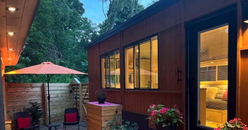 Cozy wooden tiny house at dusk with warm interior lighting, a private patio, black chairs, and a bright orange umbrella.