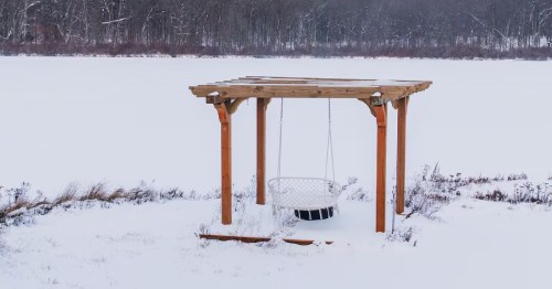 A wooden pergola with a hanging chair swing sits in a wide, snow-covered field near a winter forest.