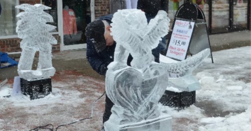 An artist carves intricate ice sculptures, including a heart with wings, outside Bailey T's clothing store during a winter festival.