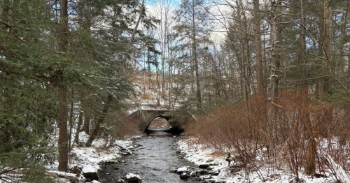 A stone arch bridge spans a rocky stream in a snow-dusted forest with evergreen and bare trees.