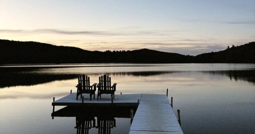 Two Adirondack chairs sit on a quiet wooden dock, overlooking a glassy lake and silhouetted mountains at twilight.