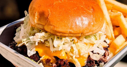 A delicious-looking burger on a metal tray being held up in someone's hand. The burger is tall and features a toasted bun, a juicy patty, melted cheese, and a generous helping of shredded lettuce. A side of crispy fries completes the scene.