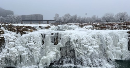 A waterfall is heavily frozen with white ice and snow, flowing into dark green water under a foggy, gray sky, with urban buildings in the background.
