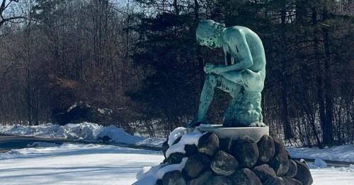Bronze statue of a pensive, seated man on a stone base in a snowy landscape.