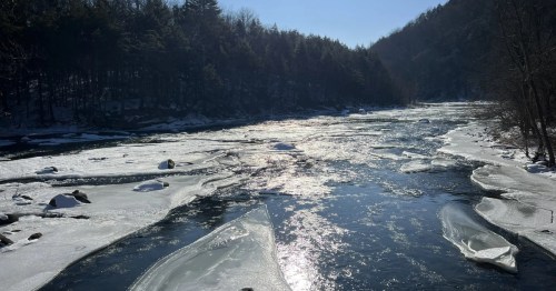 An icy river flows through a snowy forest valley under a bright, sunny sky with large ice floes on the water.