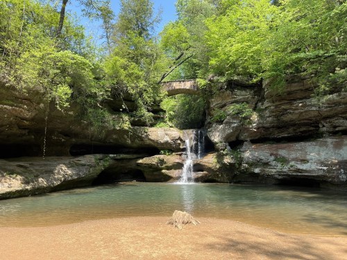 Upper Falls in Hocking Hills, Ohio, featuring a scenic waterfall cascading into a clear pool, surrounded by lush greenery, rocky cliffs, and a stone bridge above.