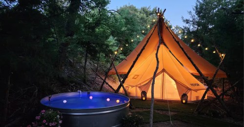 A glowing glamping tent and a blue-lit soaking tub sit tucked away in a dark forest under festive string lights.