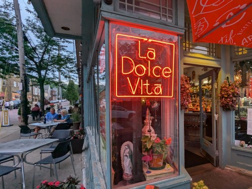 A vibrant evening street view of La Dolce Vita, a cozy Italian restaurant in Cleveland’s Little Italy. The image features a glowing red neon sign reading "La Dolce Vita" in the front window, with decorative plants and ornaments inside. The entrance is adorned with hanging flower baskets, and an open door reveals a warmly lit bar. Outside, diners enjoy meals at patio tables along a tree-lined sidewalk, contributing to the inviting and lively atmosphere of this walkable neighborhood.
