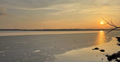 The golden light of a sunset glows over a frozen lake with a rocky shoreline in the foreground, creating a serene winter scene.
