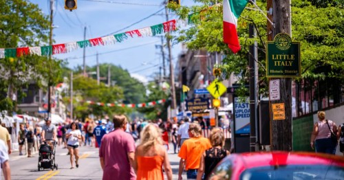 A sunlit street festival in Little Italy with crowds, Italian flags, and a historic district sign.