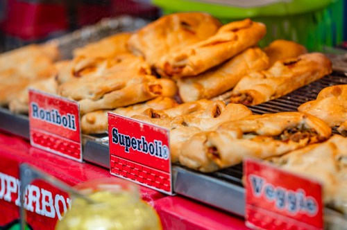 A close-up of freshly baked stuffed pastries on display at a food stand in Cleveland’s Little Italy. The golden-brown pastries, labeled as "Roniboli," "Superboli," and "Veggie," are arranged on a metal rack, showcasing their crispy crusts and savory fillings. The red signage and warm tones of the pastries create an inviting and appetizing scene, likely taken during a local festival or street fair.