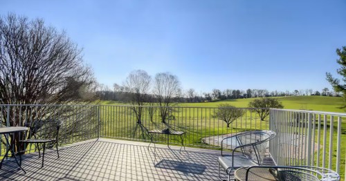 Sunlight casts long shadows across a wooden deck with metal patio furniture, overlooking a vast, rolling green field.