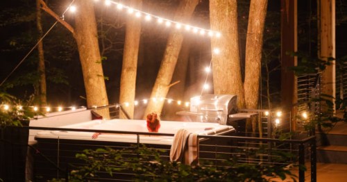 A woman relaxes in a steaming outdoor hot tub on a deck at night, illuminated by string lights in a wooded setting.