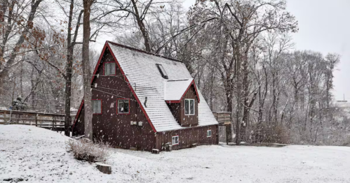 A wide shot captures The Owl's Nest A-frame cabin in Pekin, Illinois, surrounded by a snowy forest, highlighting its secluded nature setting and unique architectural design.