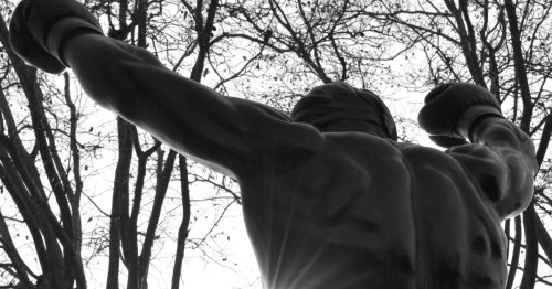 Low-angle, B&W silhouette of the Rocky statue in Philadelphia, arms raised in triumph against a backdrop of bare trees.