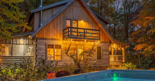 A cozy illuminated wooden cabin at twilight featuring a private balcony and a steaming outdoor hot tub.