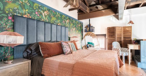 A stylish bedroom with a tall blue headboard, floral wallpaper, and rustic exposed ceiling beams.