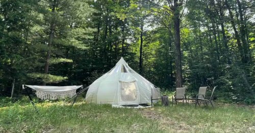 A white glamping tent with a fringed hammock and chairs set in a lush, green forest clearing.