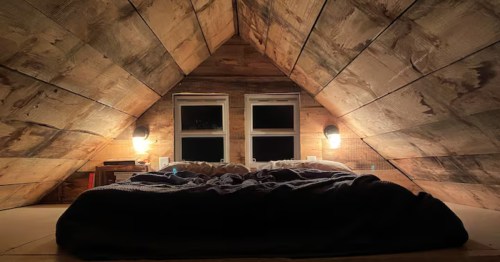 Cozy loft bedroom with a low slanted wood ceiling, warm wall lamps, and two small windows at night.