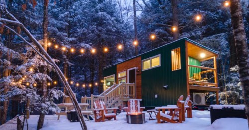 The exterior of a rectangular green tiny home, with lawn chairs around a fire pit in a snowy woodland. .