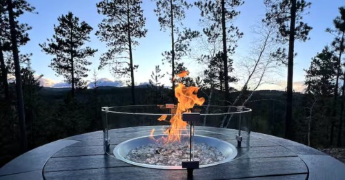 A circular outdoor gas fire pit with a glass wind guard sits on a deck overlooking a forest at twilight.