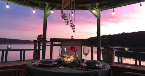 A patio at sunset, overlooking the water, with a table set with wine glasses in the foreground.