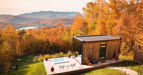 The exterior of the Luxury Glass Tiny House in Warren, Vermont. A patio with a hot tub sits next to the small, rectangular home. The home is surrounded by colorful trees and looks out on the water and mountains.