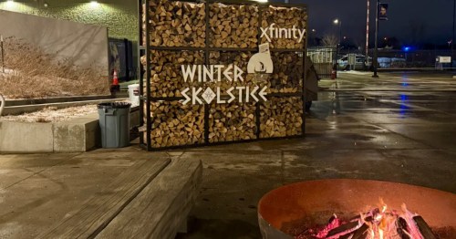 A fire burns in a metal fire pit near a wooden bench. A rack of wood in the background bears the words "Winter Skolstice" in white, stylized letters.