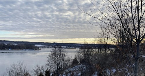 A wide, icy body of water stretching toward snow-covered hills under a dramatic, textured sky.