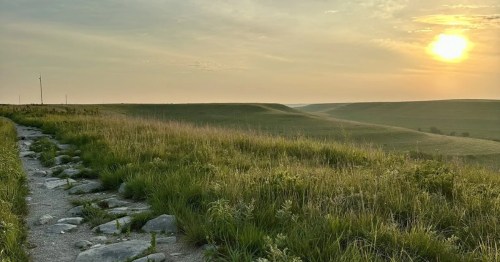 A stone-lined hiking trail winding through rolling green hills under a hazy golden sunset.