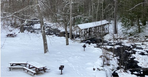 A snow-covered park features a wooden bridge over a dark stream, with picnic tables and grills dotting the white landscape.