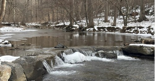 A small waterfall flows over rocks in a partially frozen river surrounded by a winter forest.