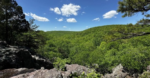 Rolling hills and rugged rock outcroppings overlook a vast, lush green valley under a soft, hazy sky.