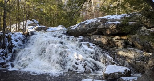 Green trees stand guard over a waterfall and rocks, all partially dusted with snow on the Rapids via Purgatory Brook Trail in New Hampshire.