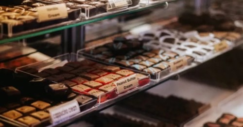 Rows of chocolates in a glass display case are arranged by flavor, including "Cappuccino Cream" and "Peanut Butter Cream". The chocolates are square-shaped with various toppings and designs, neatly presented with small flavor labels.