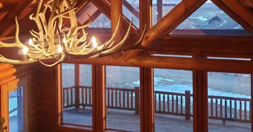 Large antler chandelier hangs in a wood cabin featuring floor-to-ceiling windows and a snowy deck view.