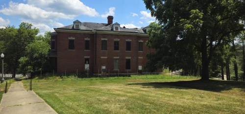 An abandoned red brick building at The Ridges in Athens, Ohio, with boarded-up windows, a deteriorating roof, and a fenced-off entrance. The structure sits among lush green trees and a well-maintained lawn under a bright blue sky with scattered clouds.