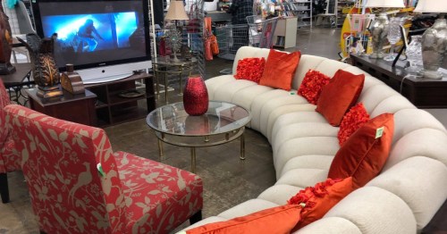 A curved white sectional sofa with orange pillows and a red patterned armchair in a thrift store setting.