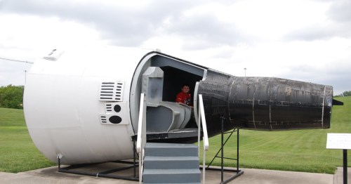 A child sits inside a large, white and black space capsule replica on a grassy field under a cloudy sky.