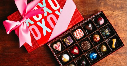 An open box of assorted artisan chocolates sits next to a red "XOXO" gift box tied with a large pink satin ribbon.