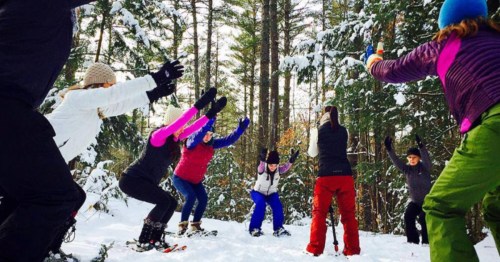 A cheerful assembly of people engaging in a snowshoe yoga, surrounded by a picturesque winter wonderland filled with snow.