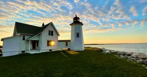 A white lighthouse and adjacent cottage on a grassy coast at sunset with a rocky shoreline.