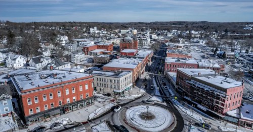 : An aerial view of a snowy town center featuring a traffic roundabout and historic brick buildings.