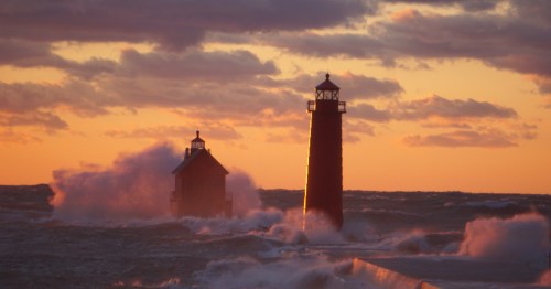 Gemini said Two red lighthouses on a pier at sunset, with large waves crashing against them under a cloudy orange sky.