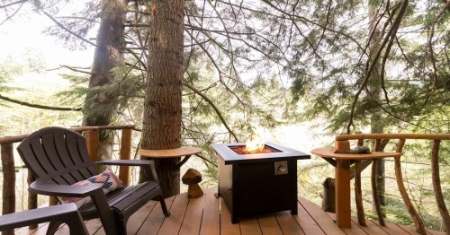 two outdoor patio chairs on a wooden deck with a fire pit in middle and trees in the background